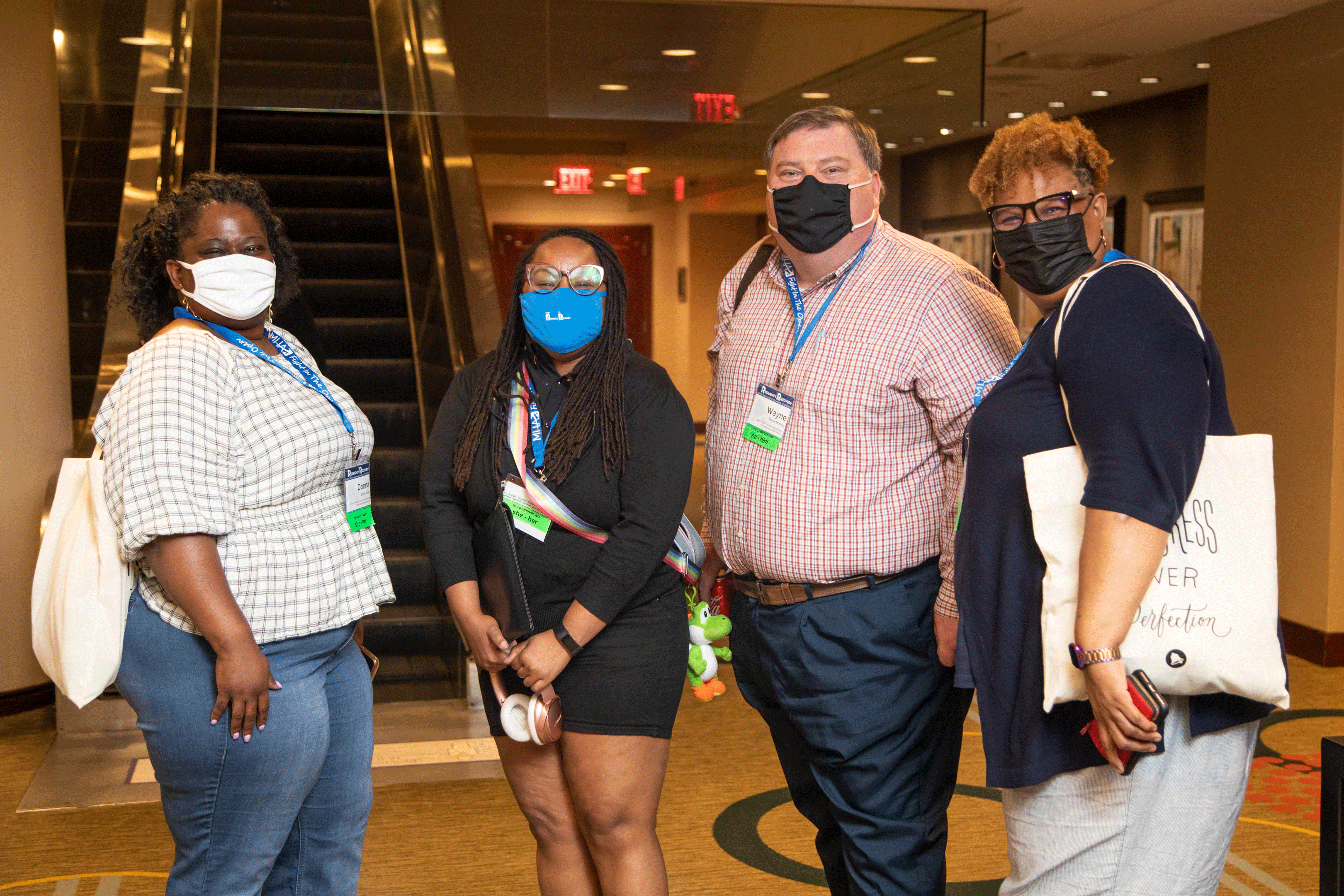4 people wearing masks and conference nametags stand together at the base of an escalator and look at the camera