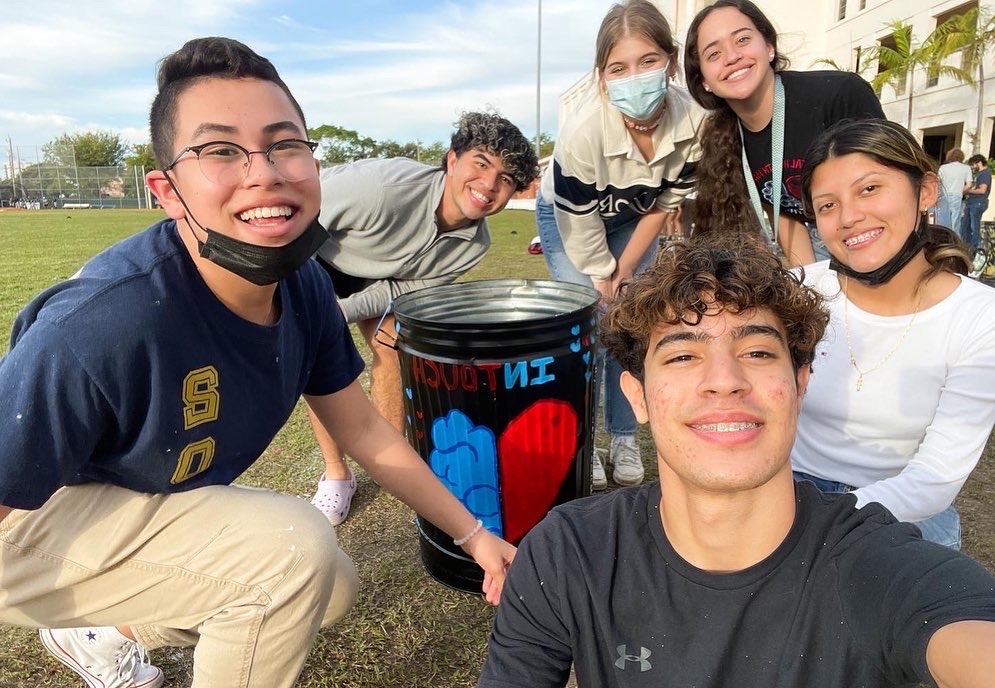 group of people stand and squat around a trash can with In Touch colorfully painted on it, smiling, some wearing face masks