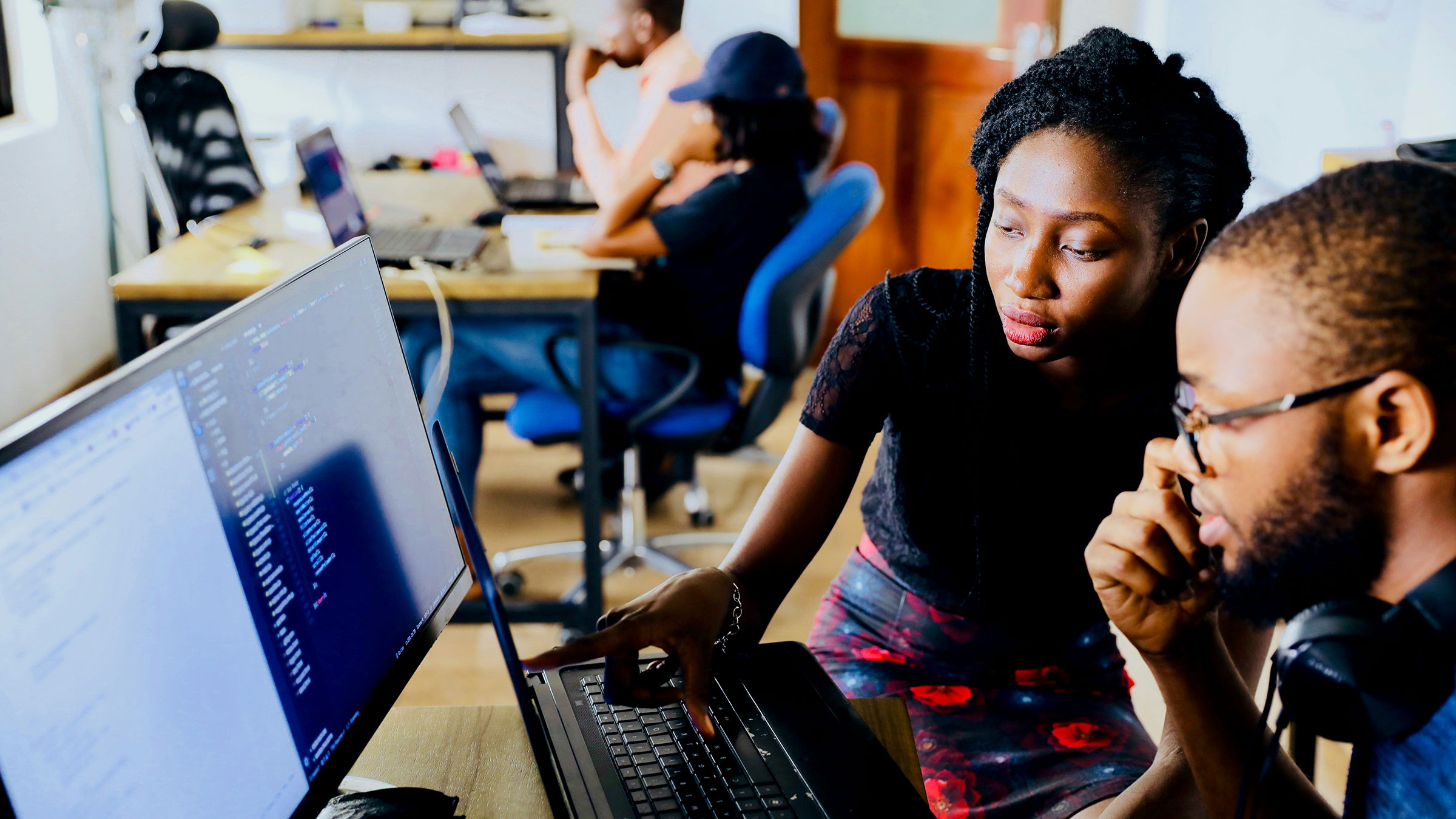 two people work at a computer together while others work at their own computers in the background