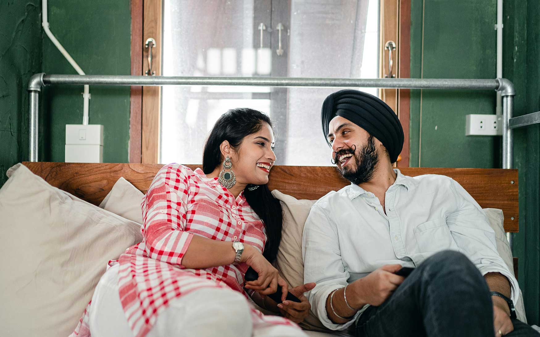 two people - one with a turban - sit on a bed talking