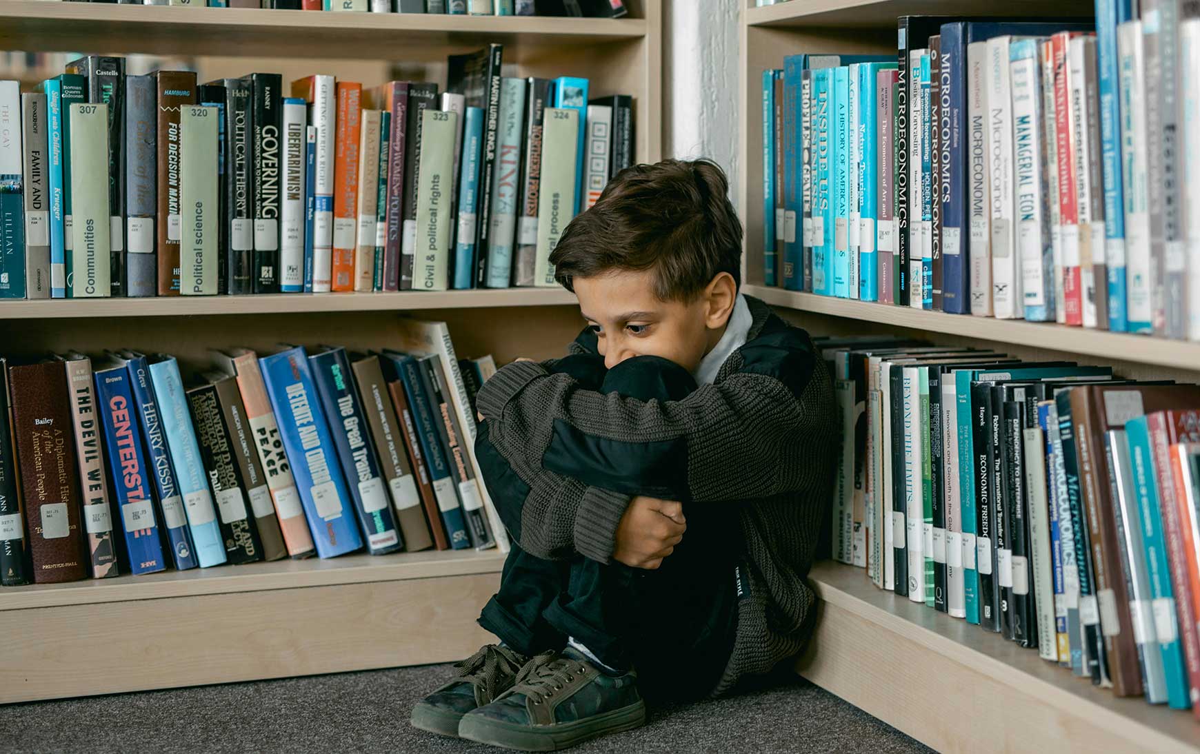 distressed child sits on floor in between bookshelves and hugs knees