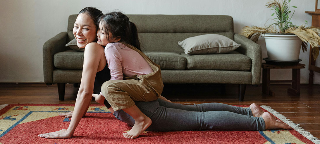person stretches out in yoga pose on floor while child sits on their back
