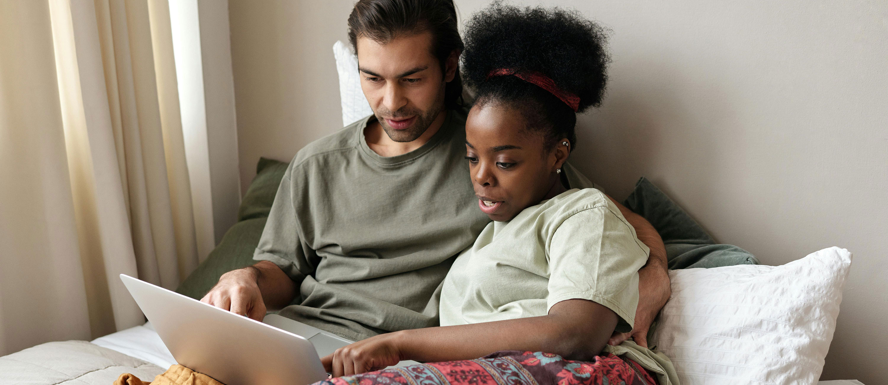 two people sit on a bed looking at a laptop