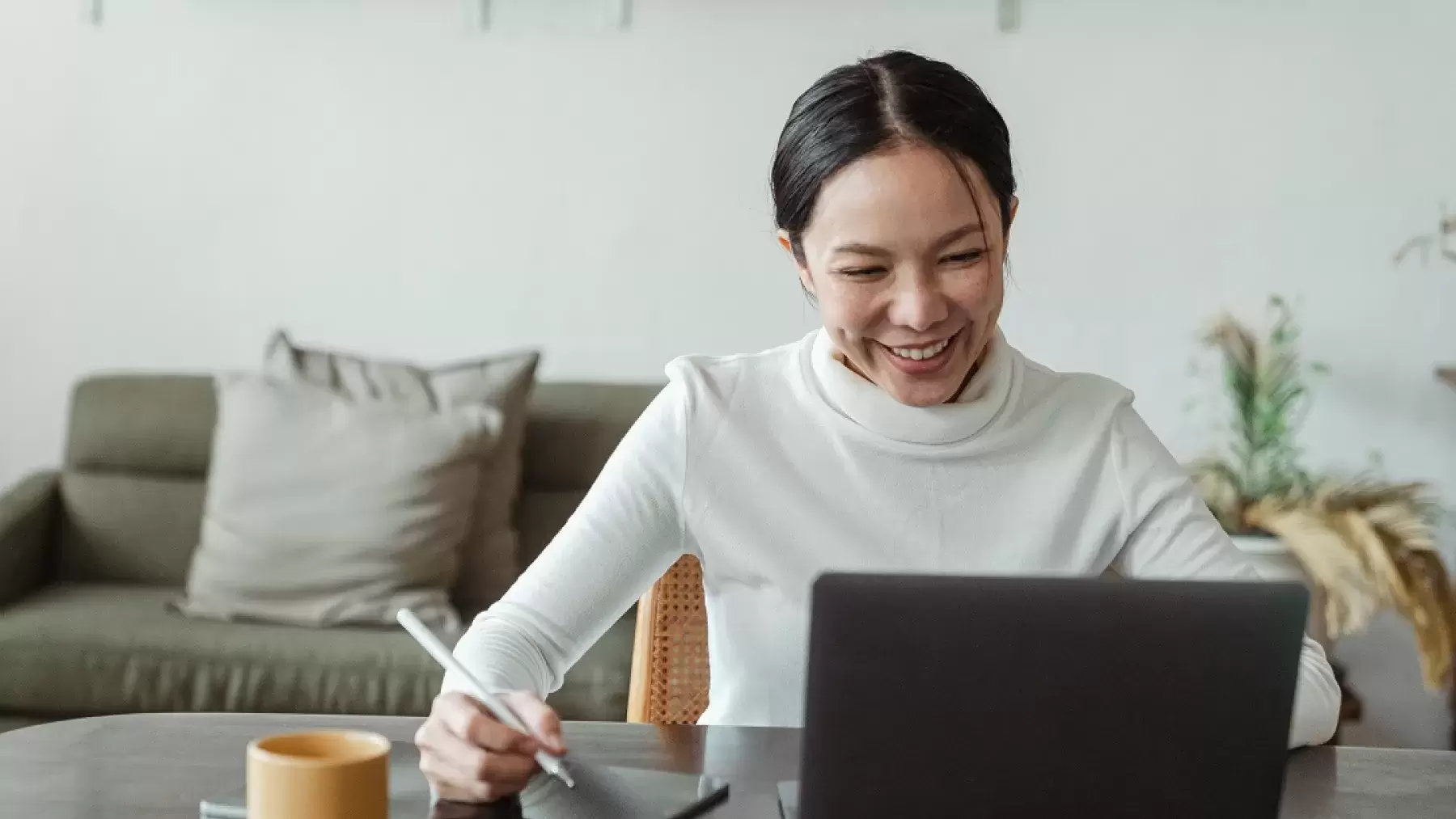 Woman with white turtleneck smiling and looking at computer screen while writing on a pad.