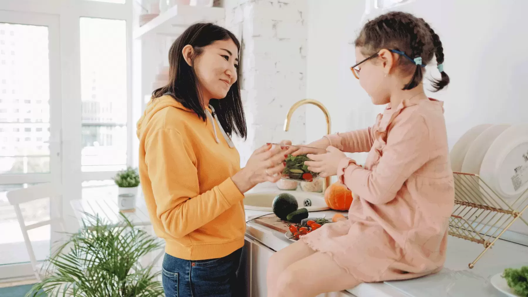 woman in orange sweatshirt is washing vegetables with little girl who is sitting on counter