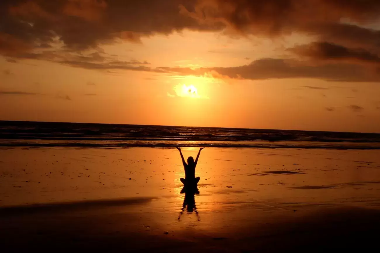 person sits on an empty beach and raises arms to sun as it rises