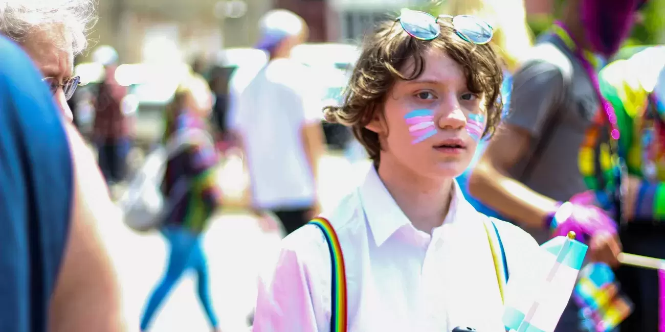 young person with transgender flag painted on cheek and wearing rainbow suspenders walks outside