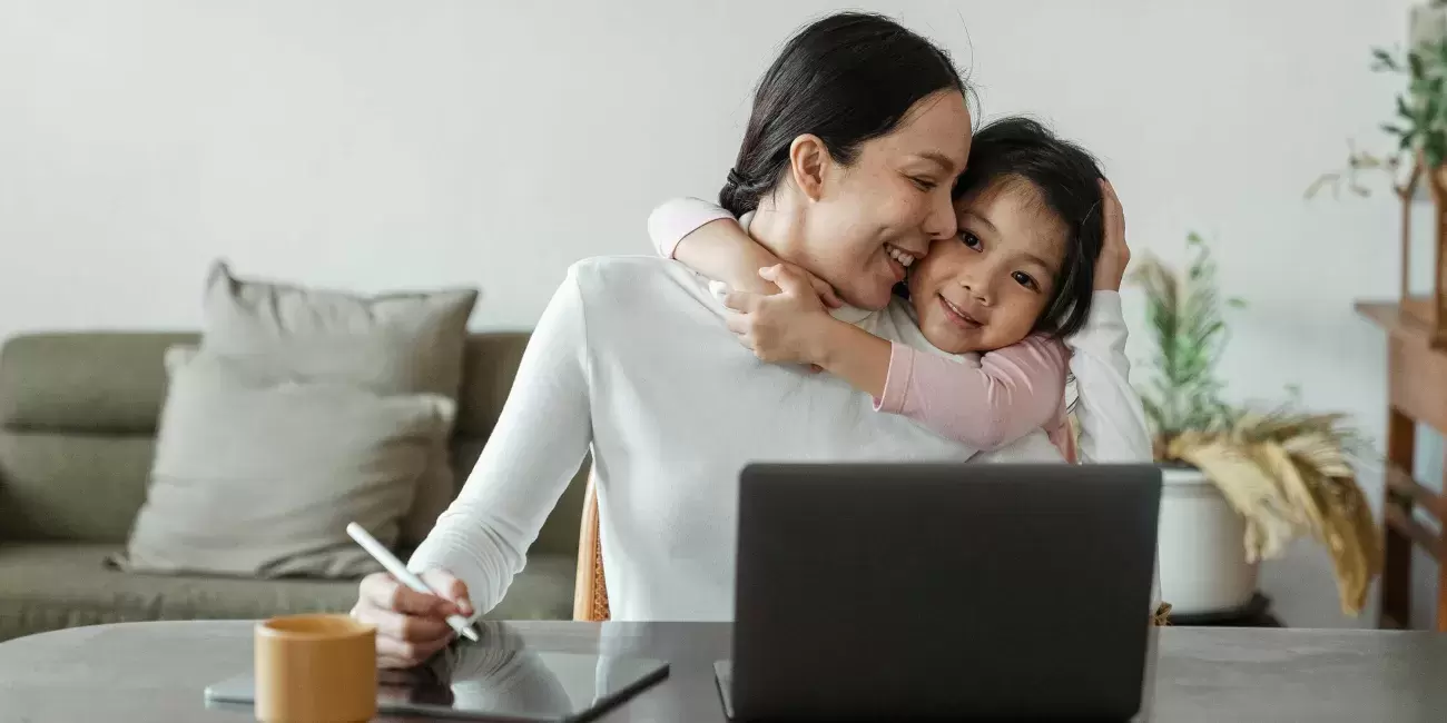 Daughter hugs mom around neck while mom works on laptop
