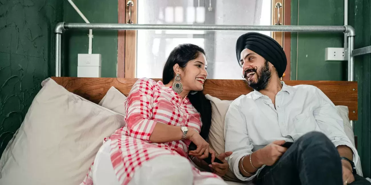 two people - one wearing a turban - sit smiling and talking in bed