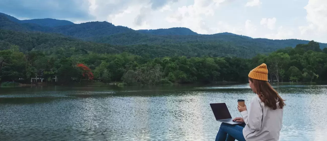 person sits with laptop by mountain lake