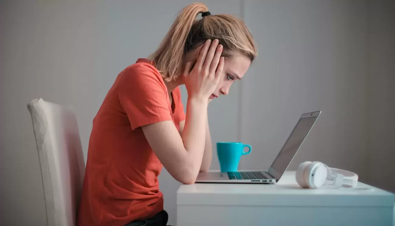 woman leans over computer at desk with hands on head, clearly stressed out