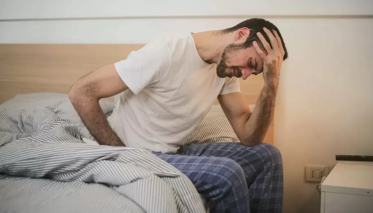 man wearing pajama pants and white t-shirt sits hunched over on side of bed with head in hands