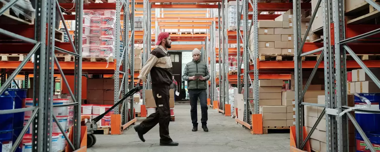 one person pulls a cart loaded with boxes in a warehouse while another checks things off on a clipboard