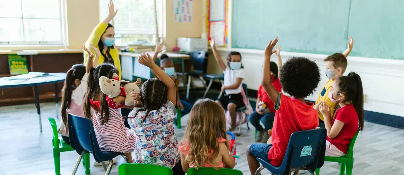 group of children in a classroom sitting in a circle with teacher and raising their hands