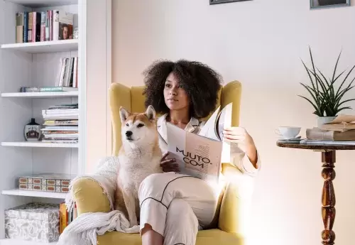 individual sits in a yellow chair reading a magazine with their dog, bookcase in background, table with plant on it to their side