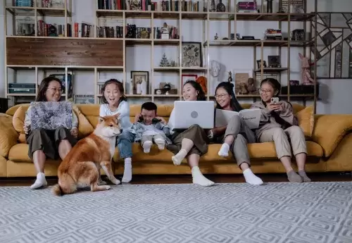 Family sits on sofa and looks at computers, books, and phone, while dog sits in front