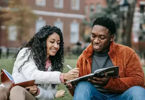 Two students of color look at books together while sitting on the grass