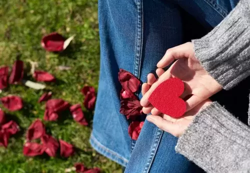 hands hold a red felt heart and there are rose petals on the grass underneath