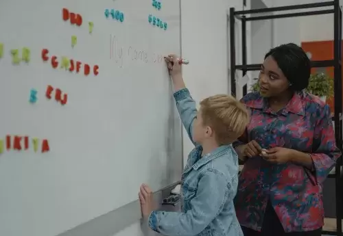 teacher stands at dry erase board while student writes on it