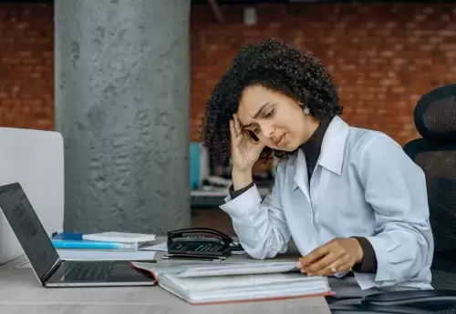 stressed person sits at desk with head in hand and laptop and notebooks in front of them