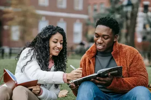Two students of color look at books together while sitting on the grass