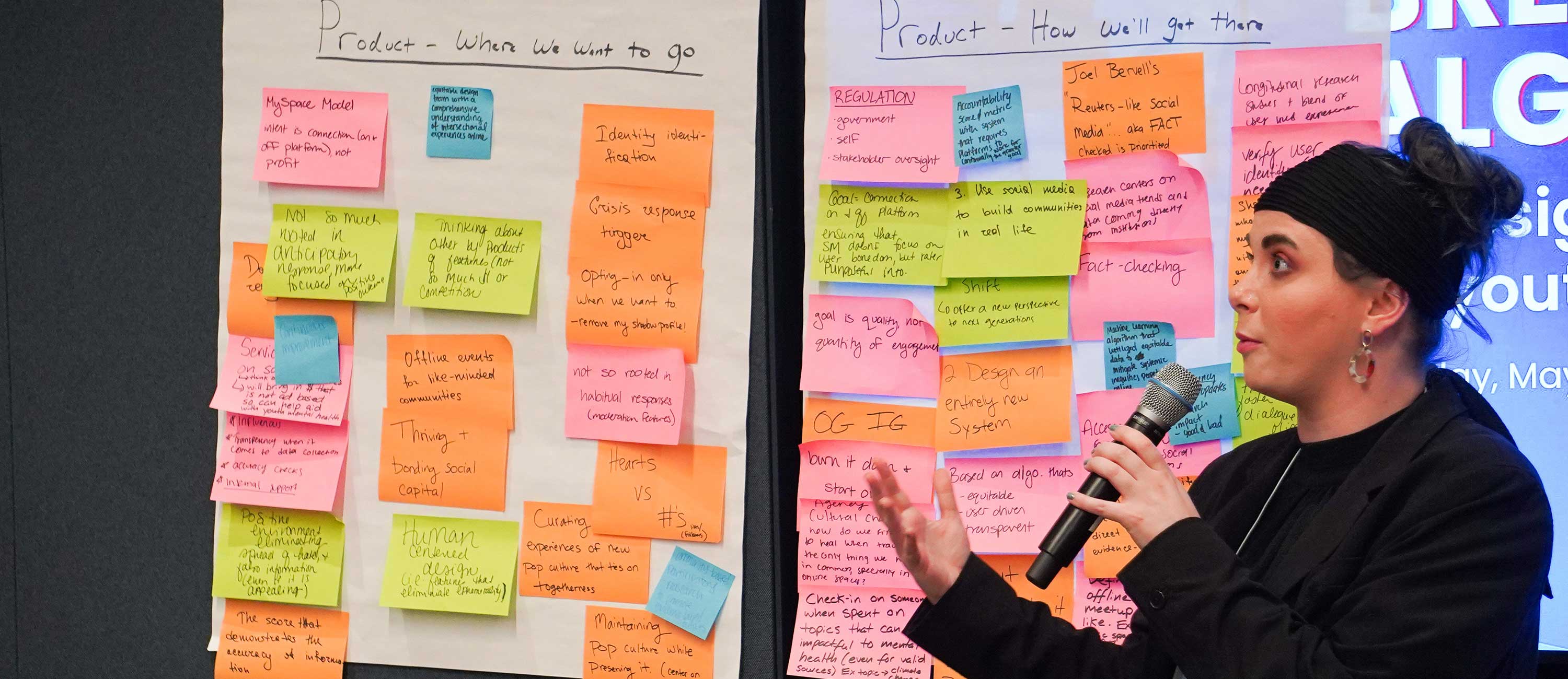 person stands in front of posterboards covered in sticky notes while speaking into microphone