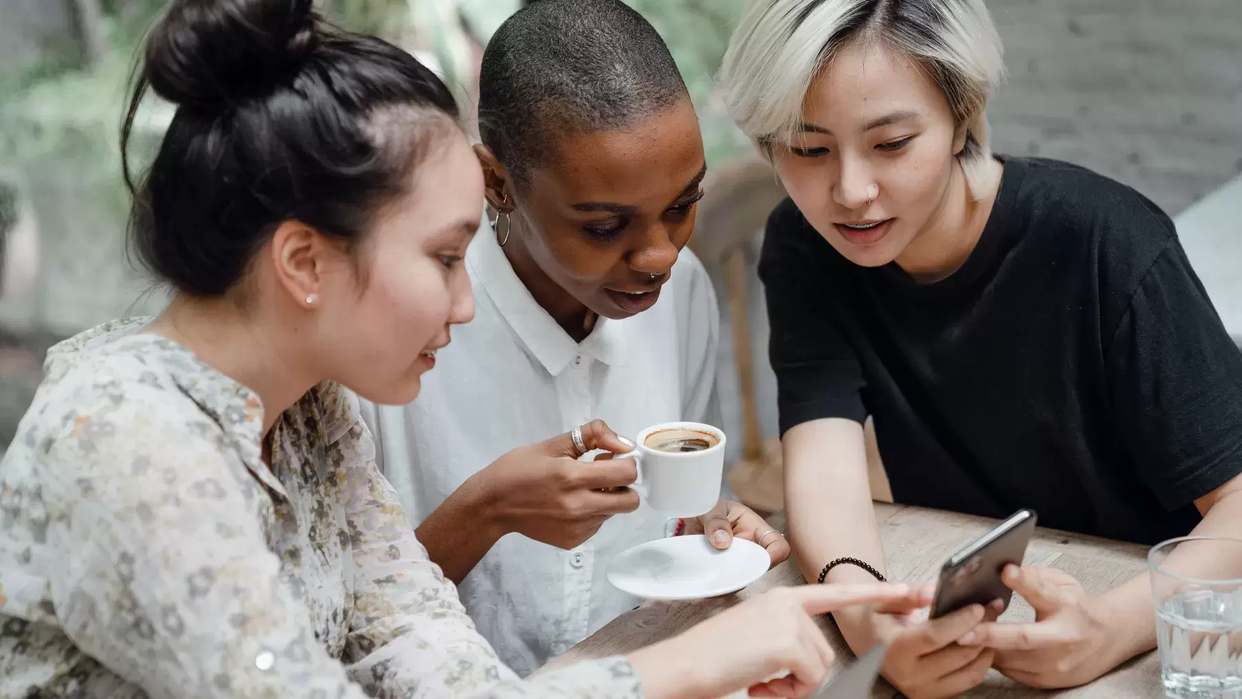 3 people are huddled together looking at a cell phone. 1 person is holding a cup of coffee