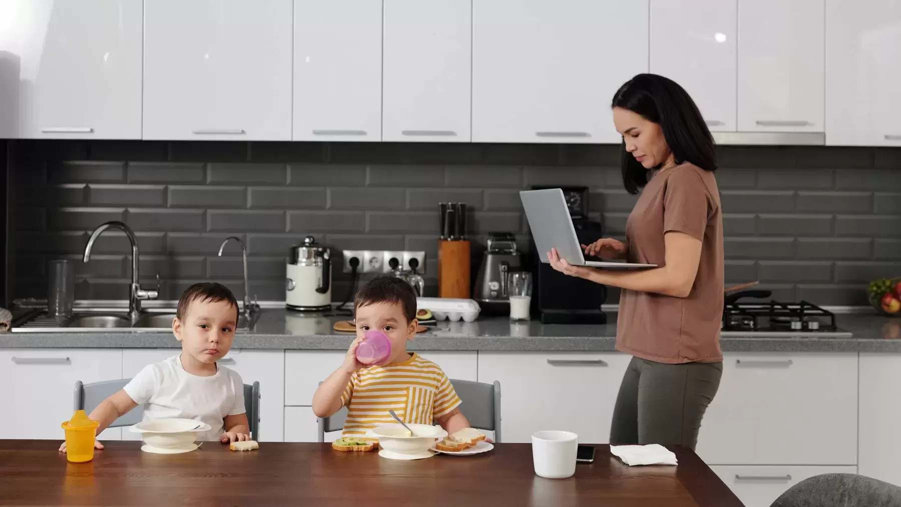 person is on laptop in kitchen standing behind two kids eating at a table