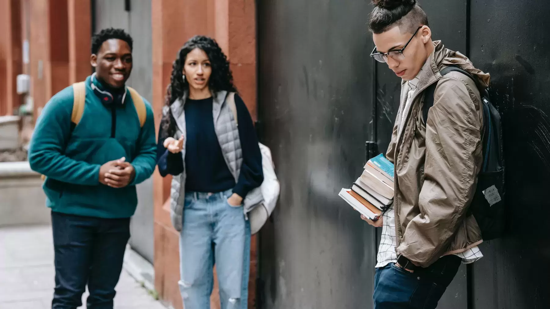 1 young person is standing, holding books, and looking down while 2 young people are looking at him from a distance