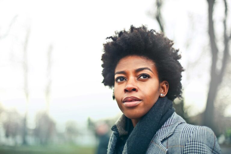 person wearing scarf and coat stands outdoors with trees behind them