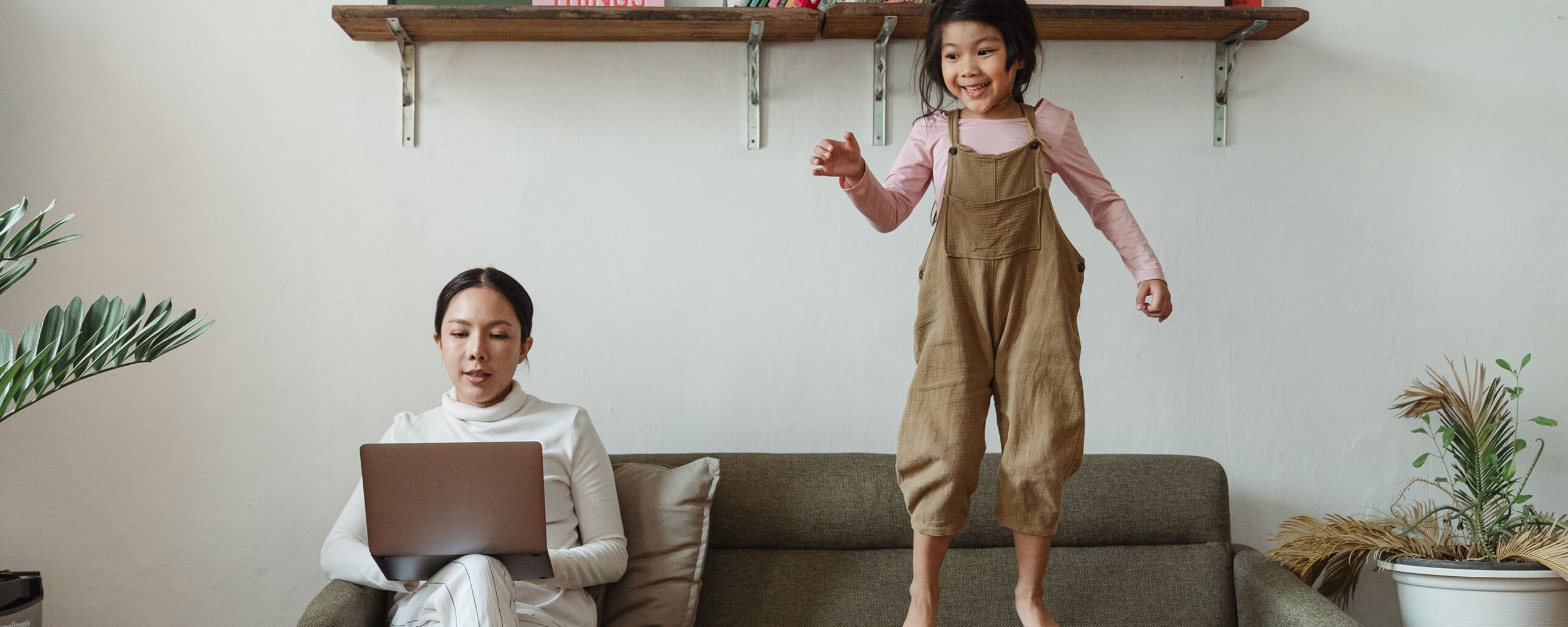 person sitting on couch works on laptop while child jumps next to them