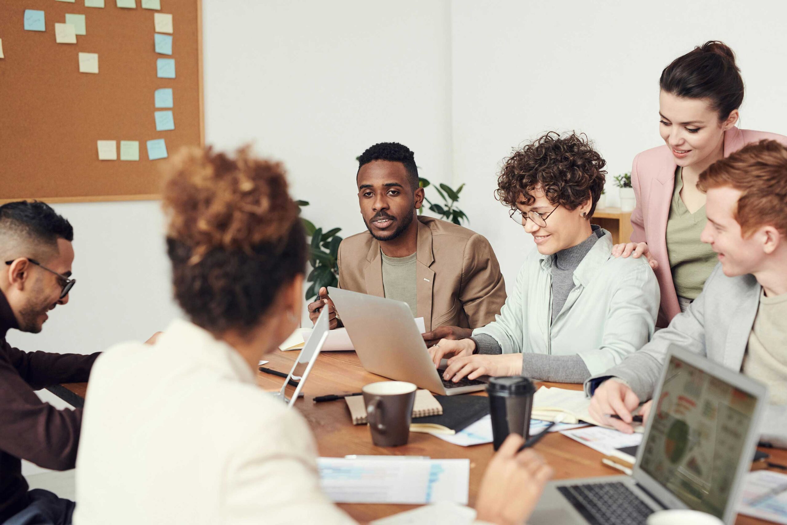people sit and stand around a conference table with computers open in front of them