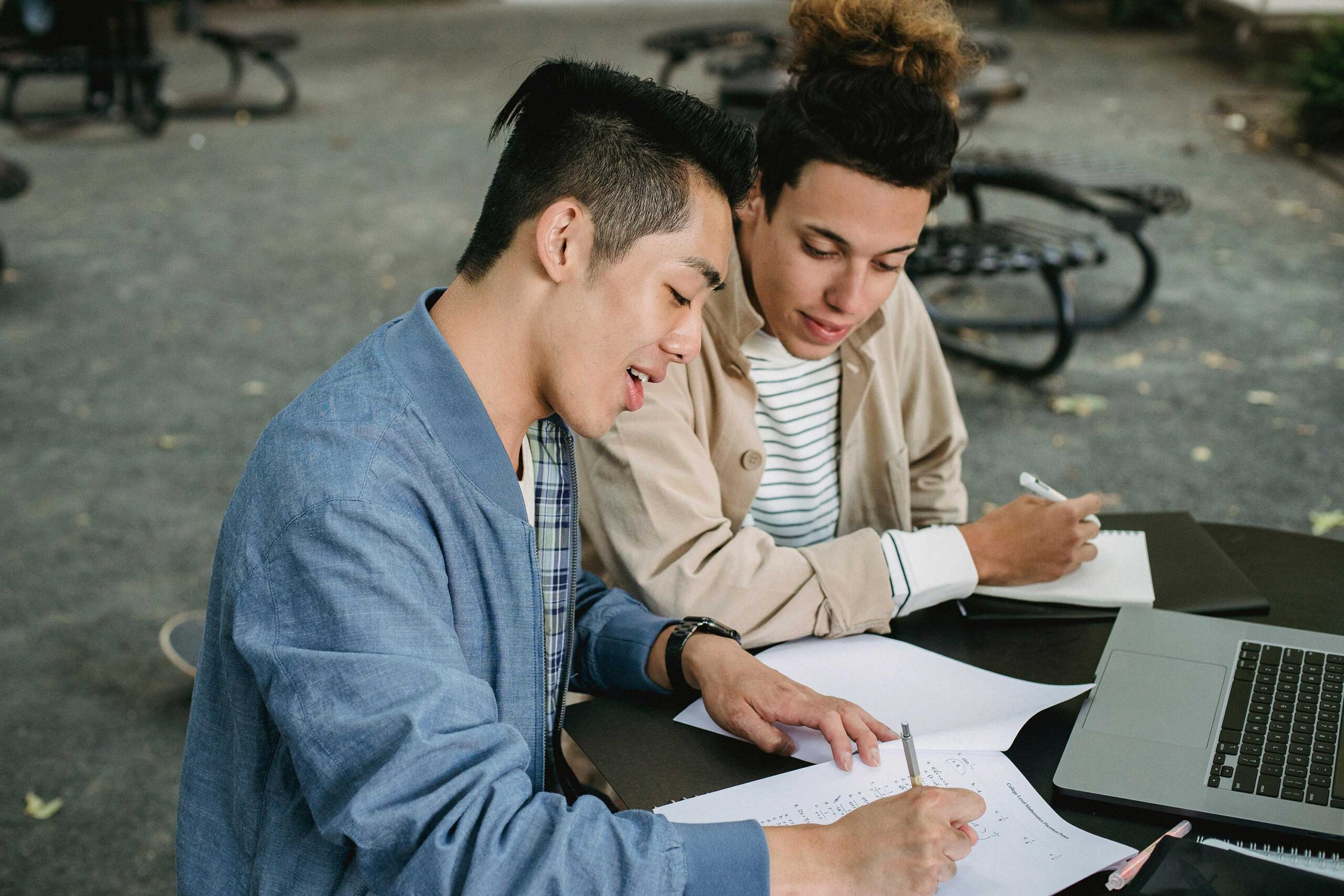 two people sit next to each other at a table writing on paper with laptop open in front of them