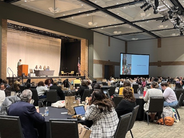 People sit at tables in an auditorium listening to a panel of people who are presenting on stage