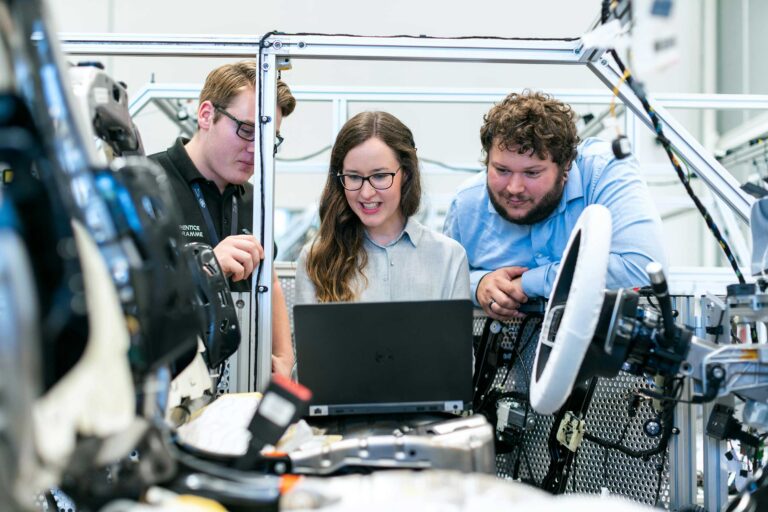 3 people stand in a workshop and look at a computer with other equipment around them